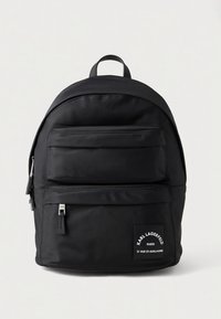 Μαύρο σακίδιο πλάτης από νάιλον με μπροστινή τσέπη με φερμουάρ, ρυθμιζόμενους ιμάντες και διακοσμητική ετικέτα με λογότυπο. Λεία υφή με κυκλικό σχήμα και μινιμαλιστικό σχέδιο.