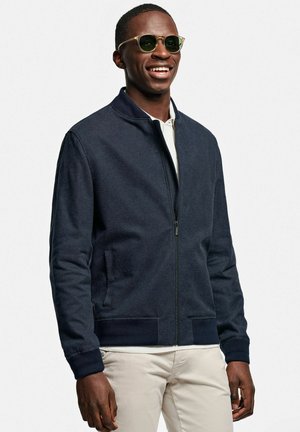 Lachende man met groene ronde zonnebril, marineblauw jack met rits, wit shirt eronder en lichtbeige broek tegen een effen witte achtergrond.