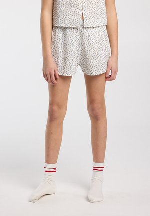 Beine einer Person, die weiße Shorts mit Blumenmuster, ein passendes ärmelloses Oberteil und weiße Socken mit roten Streifen trägt, stehend auf weißem Hintergrund.