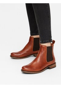 Botas de tobillo de cuero marrón con paneles laterales elásticos oscuros, punta redonda y tacón apilado. La superficie es suave con un brillo sutil.