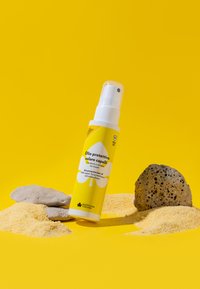Spray solare in una bottiglia bianca e gialla su uno sfondo giallo texturizzato; circondata da pietre grigie e granuli gialli simili alla sabbia.