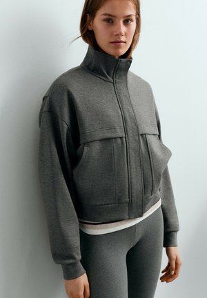 Jeune femme portant une veste grise zippée avec un col montant et des leggings assortis, debout contre un mur de couleur claire.