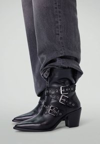 Bottes noires en cuir à bout pointu, avec des sangles à boucle en argent et un talon épais. Portées avec un pantalon en denim gris retroussé.