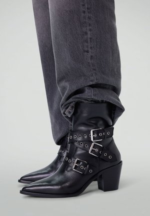 Zwarte leren enkellaarsjes met puntige neuzen, zilveren gespbandjes en een chunky hak. Draag ze bij opgerolde grijze denim broek.