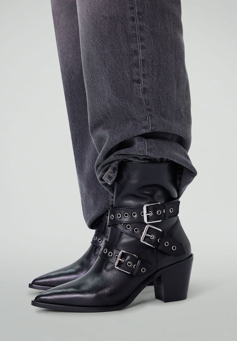 Bottes noires en cuir à bout pointu, avec des sangles à boucle en argent et un talon épais. Portées avec un pantalon en denim gris retroussé.