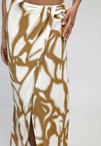 Jupe longue en tissu blanc avec des motifs abstraits marron et crème, détail fente haute et design de nœud torsadé à la taille.