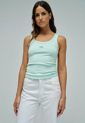 Femme portant un débardeur côtelé bleu clair avec le texte "SALSA JEANS" et un pantalon blanc, debout devant un fond gris clair uni.