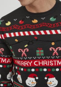 Svart strikkegenser med juletitler, inkludert snømenn, sukkerstokker og ornamenter, med røde og grønne detaljer og et bånd med teksten "GOD JUL".
