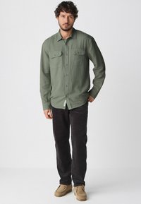 Grünes Hemd mit Knöpfen und zwei Brusttaschen, kombiniert mit schwarzen Cordhosen und beigefarbenen Schuhen. Weicher Stoff, lässiges Design und gerade Passform.