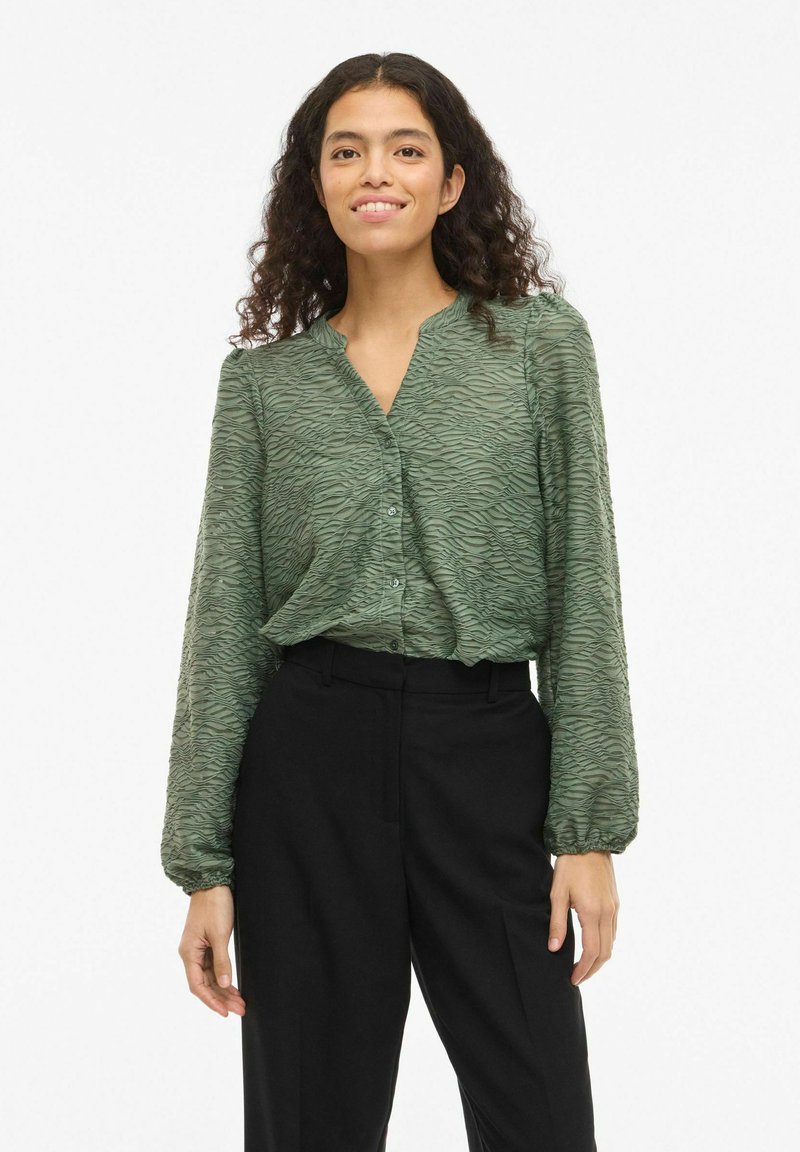 Blusa verde texturizada con escote en V y cierre de botones, con mangas largas recogidas. Combinada con pantalones negros a medida.
