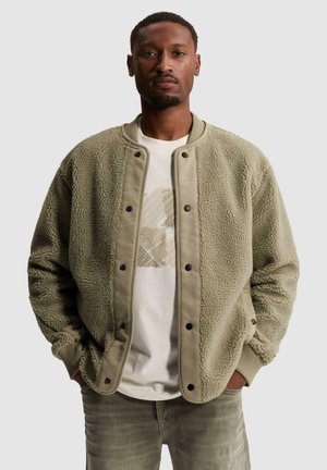 Homme portant une veste polaire beige sur un t-shirt graphique blanc, les mains dans les poches, debout devant un fond clair uni.