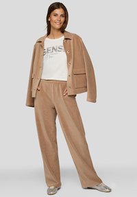 Frau steht und trägt eine beige Cordjacke und weit geschnittene Hosen mit einem weißen T-Shirt mit Grafik und silbernen Turnschuhen vor grauem Hintergrund.