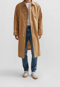 Lätt, beige trenchcoat med en stor krage, knappstängningar och sidofickor, bärs över en vit T-shirt och blå jeans.