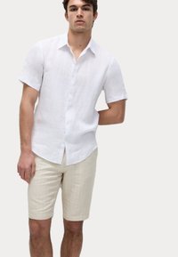 Camisa de lino blanca de manga corta con botones al frente, combinada con pantalones cortos beige con rayas finas, que cuentan con un ajuste relajado y una textura suave.