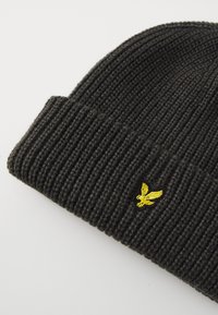 Gorro de punto acanalado negro con puño plegable y un pequeño logotipo de águila bordado en amarillo en la parte frontal. Tejido texturizado para abrigo.