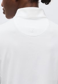 Κοντινό πλάνο ενός ατόμου που φοράει λευκό ριμπ μπλουζάκι με ένα μικρό, διακριτικό λογότυπο σχήματος ζιγκ-ζαγκ στο πίσω μέρος του γιακα.