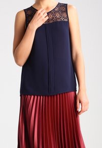 Haut sans manches bleu marine avec un empiècement en dentelle aux épaules, associé à une jupe bordeaux plissée. Tissu lisse avec des textures et des couleurs contrastées.