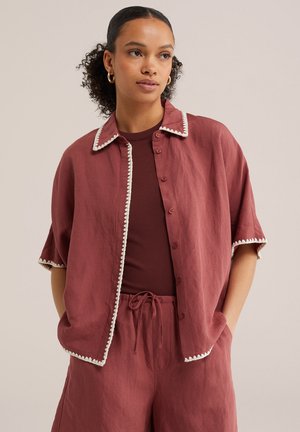 Žena nosi voľnú bordovú košeľu na gombíky a zodpovedajúce nohavice so šnúrkou, s bielym lemovaním na okrajoch košele a zlatými kruhovými náušnicami.