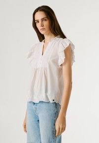 Νεαρή γυναίκα που φοράει λευκό αμάνικο τοπ με βολάν και λεπτομέρειες με οπές, και ανοιχτό γαλάζιο high-waisted τζιν, στέκεται μπροστά σε απλό φόντο.