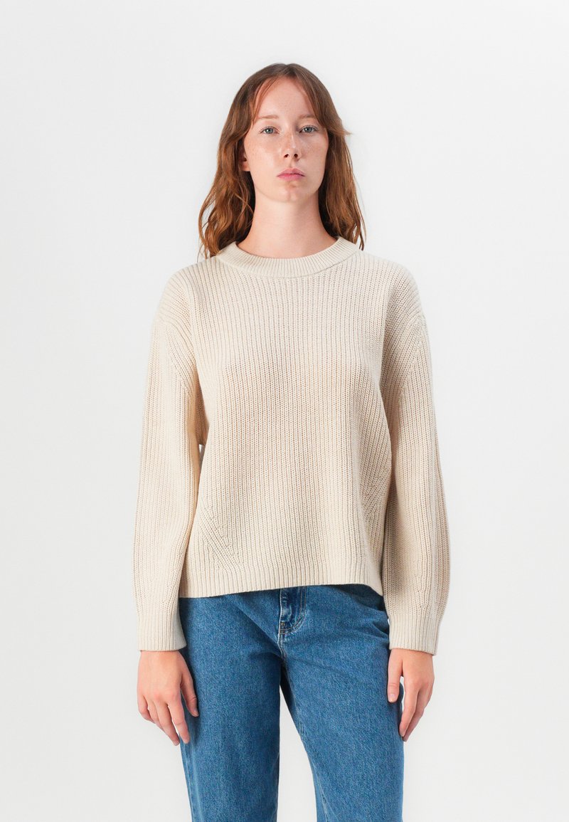 Pull en tricot beige avec un col rond, texture côtelée et manches longues, associé à un jean bleu clair sur un fond neutre.