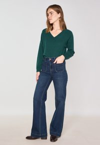 Maglione verde a V con maniche lunghe abbinato a jeans a vita alta svasati di colore blu scuro. Calzature marroni e cuciture dettagliate sui jeans.