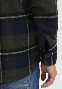 Groene en marineblauwe geruite flanellen shirt met een knoopmanschette; heeft witte strepen en een gestructureerde stof, gedragen over een spijkerbroek.