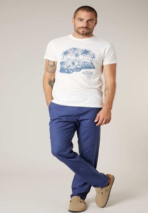 Homme aux cheveux courts et tatouages porte un T-shirt blanc avec un motif de la Riviera italienne, un pantalon bleu et des chaussures beige sans lacets, debout de manière décontractée avec les jambes croisées.