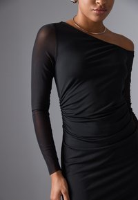 Robe noire avec de longues manches transparentes et un design froncé à la taille, dotée d'un décolleté bateau et d'une texture lisse.