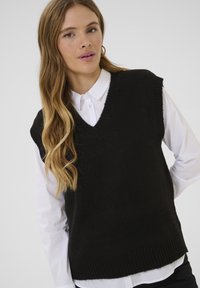 Black knitted sleeveless sweater with a V-neck, ribbed hem, textured fabric, layered over a white collared shirt. No visible patterns or accents.