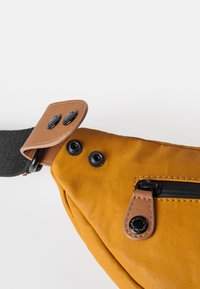 Tan stoffen schoudertas met een gestructureerd oppervlak, uitgerust met een zwarte rits en stevige hardwareaccenten. Inclusief twee zwarte drukknoopsluitingen.