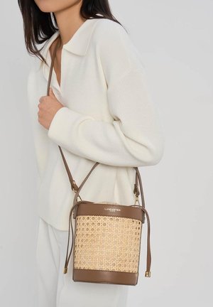 Vrouw die een witte top en broek draagt en een bruine en beige geweven boodschappentas met schouderriem vasthoudt, merk Lancaster zichtbaar.