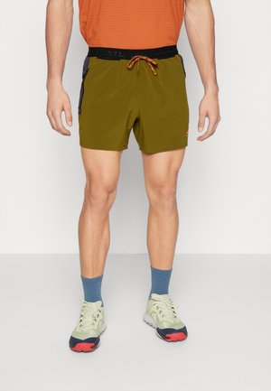 Mann trägt olivgrüne Sportshorts mit schwarzen Seitentaschen, orangefarbenem Kordelzug, blauen Socken und hellgrünen Laufschuhen.