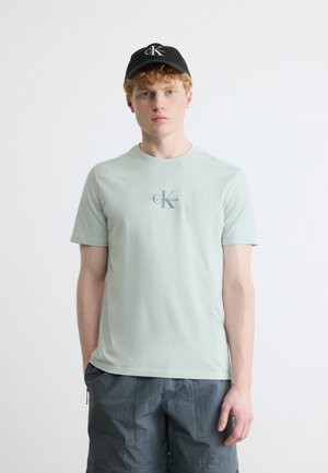 Junger Mann mit lockigem roten Haar, der ein hellgrünes Calvin Klein T-Shirt, dunkel strukturierte Shorts und eine schwarze Calvin Klein Kappe trägt.