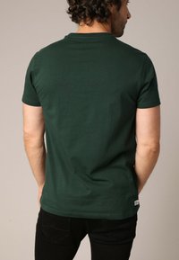 Korte mouwen, donkergroene t-shirt van katoen met een ronde hals. Heeft een rechte zoom en een klein merklabel aan de onderste linkerzijde.