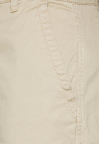 Pantalons en mélange de coton beige présentant une poche avant, des coutures visibles et une texture lisse. Le tissu a un léger éclat.