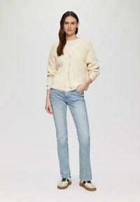 Cardigan chiaro con bottoni, lavorato a maglia strutturata, indossato sopra una maglietta a righe. Jeans blu e sneakers multicolori completano l'outfit.