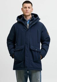 Marineblå isoleret parka med to frontlommer, lynlås- og knaplukning, justerbar hætte og glat tekstur. Lagdelt over en denimskjorte.