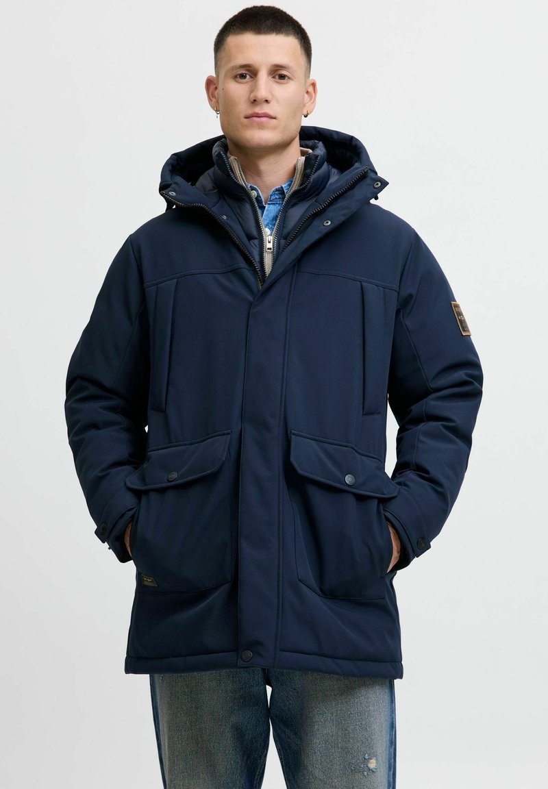 Marineblå isoleret parka med to frontlommer, lynlås- og knaplukning, justerbar hætte og glat tekstur. Lagdelt over en denimskjorte.