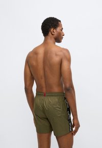 Oliivrohelised ujumispüksid, millel on elastne vöökoht ja tagatasku. Omavad musti küljeribasid logoga, siledat tekstuuri ja kerget materjali.