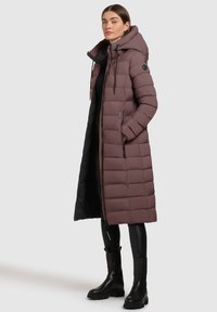 Polstret lang frakke i mauve med hætte, med quilted sektioner, to lynlåsede lommer og en sort indvendig foring. Parret med sorte støvler.