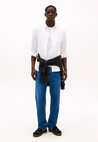 Homem vestindo camisa branca de botões sobre t-shirt branca, jeans azuis, ténis pretos e suéter preto amarrado à cintura, em pé sobre fundo branco.