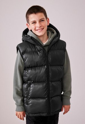 Gilet imbottito nero con chiusura a zip, cappuccio e tasche laterali, indossato sopra un maglione verde testurizzato con scollatura rotonda.