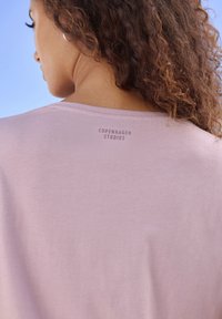 Hellrosa T-Shirt aus glattem Stoff, mit rundem Halsausschnitt und dem Markennamen "COPENHAGEN STUDIOS", der nahe dem Kragen aufgedruckt ist.