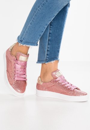 Person iført blå afkortede jeans og glitrende pink sneakers med skinnende satin snørebånd på en hvid gulvbaggrund.