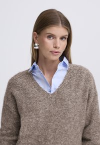 Svetlo rjav v-izrez pulover z mehko teksturo čez svetlo modro srajco z ovratnikom. Model nosi velike srebrne uhane in prikazuje naravne lase.