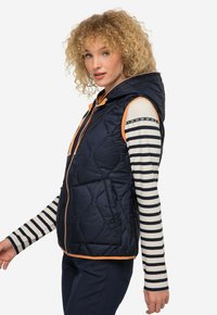 Gilet imbottito blu navy con cappuccio, dettagli in arancione sulla zip e due tasche frontali. Indossato sopra una maglietta a maniche lunghe a righe, che mostra un look a strati.