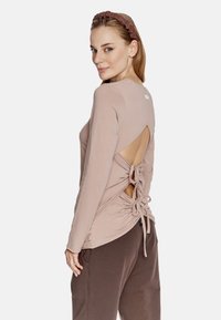 Beżowa bluzka z długim rękawem i otwartych plecach, z regulowanymi sznurkami i miękkiego materiału. Noszona z ciemnobrązowymi spodniami, włosy w brązowej opasce.