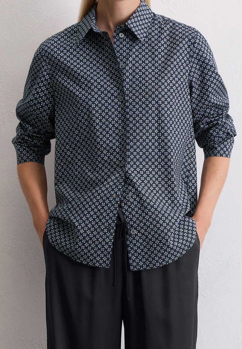 Personne portant une chemise noire à motifs avec boutons, manches retroussées et pantalon noir ample, debout les mains dans les poches.