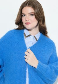 Eine Frau mit welligem braunen Haar trägt eine blaue, flauschige Strickjacke über einem blau gestreiften Hemd mit Leopardenmuster am Kragen und hält die Strickjacke geschlossen.