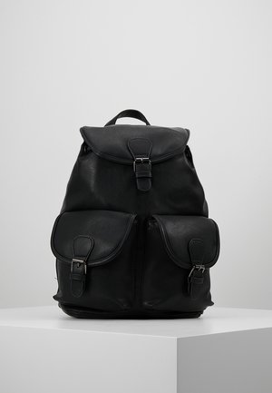 Schwarzer Leder-Rucksack mit oberer Klappe und zwei vorderen Taschen mit Schnallen auf einem weißen Podest vor einem schlichten hellgrauen Hintergrund.